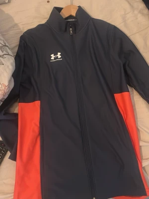 Blå och röd vindjacka från Under Armour - Sportig vindjacka från Under Armour i mörkblått med röda paneler på sidorna. Jackan har hel dragkedja framtill, hög krage och logga på bröstet. Tillverkad i lätt polyester som passar perfekt för aktiva dagar. Byxor till hoodien ingår 