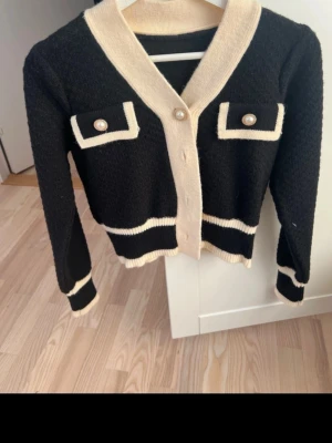 Svart kofta med vita detaljer - Stilren svart kofta med kontrasterande vita kanter och dekorativa fickor framtill. Koftan har pärlknappar och ribbade muddar vid ärmslut och nederkant. Perfekt för dig som gillar en chic och klassisk look.