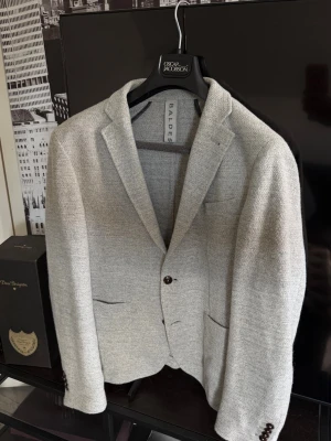 Ljusgrå kavaj från Baldesarini - Stilren ljusgrå kavaj från Baldesarini med två knappar framtill, klassisk krage och tre fickor. Kavajen har en strukturerad yta och bruna knappar vid ärmsluten. Perfekt för en dressad look.