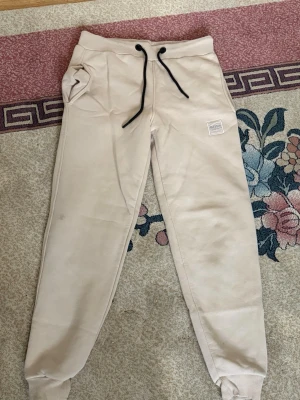 Beige mjukisbyxor från Hugo Boss - Snygga beige mjukisbyxor från Hugo Boss med svart dragsko i midjan och ribbade muddar vid bensluten. Byxorna har två sidofickor och en diskret patch med BOSS-loggan på vänster ben. Perfekta för en chill och avslappnad stil.