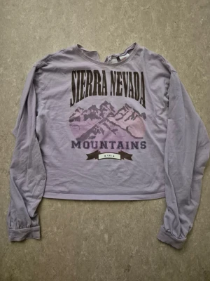 Tröja - Lila långärmad tröja med tryck av berg och texten 'Sierra Nevada Mountains' på framsidan. Tröjan har en avslappnad passform, rund halsringning med liten slits och ribbade muddar vid ärmsluten. Perfekt för en chill och casual stil.