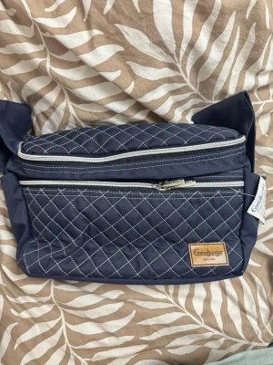 Blå organiserarväska från Emmaljunga - Snygg organiserarväska i marinblått från Emmaljunga med quiltat mönster och vita sömmar. Väskan har två dragkedjefack, justerbar rem och en liten läderdetalj med logga. Perfekt för att hålla ordning på småprylar och accessoarer.