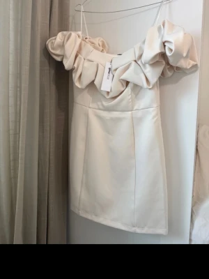 Krämvit kort klänning Gina Tricot - Supersnygg kort klänning från Gina Tricot i krämvit nyans. Klänningen har stora volangdetaljer vid axlarna och en axelbandslös design som ger en trendig look. Tillverkad i ett slätt, något glansigt material som känns lyxigt.