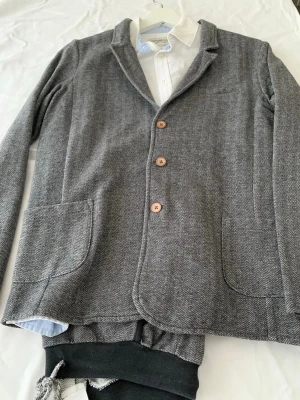 Grå kostym med kavaj och byxa till barn 12-14 åring  - Lindex kavaj för pojke i storlek 158-164. Kavajen är grå och tillverkad i bomull. Modellen har knäppning framtill, utanpåfickor och enkel krage. Passar till vardag och mer uppklädda tillfällen för barn i åldern cirka 12–14 år beroende på längd.   Ingår även: • Vit skjorta i storlek 158 som passar bra till  