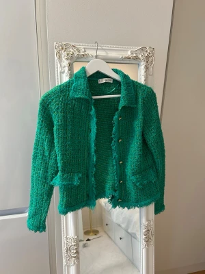 Grön bouclékofta från MNG - Snygg grön bouclékofta från MNG med dekorativa knappar och två fickor framtill. Koftan har en klassisk krage och råa kanter för en trendig look. Perfekt att slänga över en t-shirt eller klänning för extra stilpoäng.