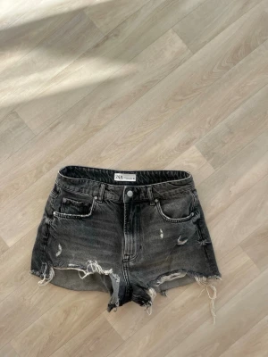 Svarta slitna jeansshorts från Zara - Säljer ett par svarta jeansshorts från Zara med råa, fransiga kanter och slitningar framtill. Shortsen har fem fickor, knappgylf och en klassisk midja. Perfekta för en avslappnad och trendig look.