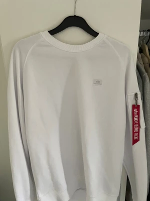 Vit sweatshirt Alpha Industries - Säljer en stilren vit sweatshirt från Alpha Industries med rund halsringning och lång ärm. Tröjan har en liten logopatch på bröstet och en ficka med dragkedja samt röd tag på ena ärmen. Perfekt för en clean och modern look.