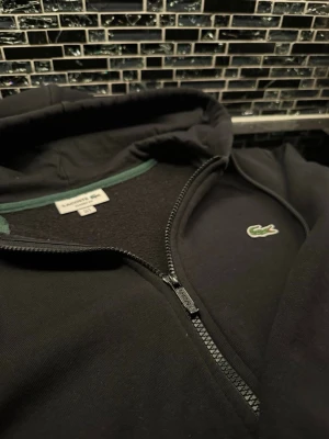 Lacoste hoodie med dragkedja  - Svart hoodie från Lacoste med dragkedja. Hoodien har huva med mjukt foder på insidan. Perfekt för en chill och stilren look. Materialet är bomullsmix som känns skönt mot huden.
