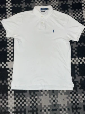 Vit pikétröja från Polo Ralph Lauren - Klassisk vit pikétröja från Polo Ralph Lauren med blå broderad logga på bröstet. Tröjan har korta ärmar, krage och knäppning med två knappar. Slim fit-modell i bomull som passar perfekt till jeans eller shorts.