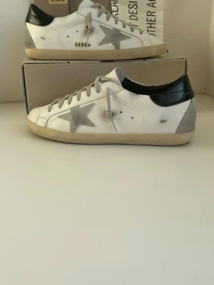 Golden Goose vita sneakers med stjärna - Snygga vita sneakers från Golden Goose med grå stjärna på sidan och svart hälparti. Skorna har gråa skosnören och en slitstark gummisula.