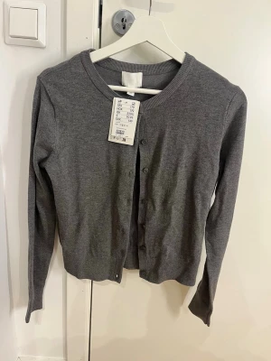 Grå kofta från H&M med knappar - Snygg grå kofta från H&M med rund halsringning och knäppning framtill. Tillverkad i mjukt, tunt material och har långa ärmar samt ribbade muddar vid hals och ärmslut. Perfekt att slänga över en topp för en enkel och clean look. Aldrig använd med prislappen kvar - nypris: 179 kr