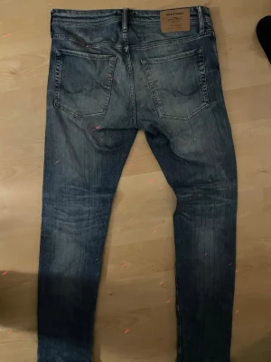 Blå slim fit jeans från Jack & Jones - Säljer ett par blå slim fit jeans från Jack & Jones med klassisk femficksdesign och snygga slitningar. Jeansen har normal midja, smala ben och är tillverkade i denim med lätt stretch för skön passform. Perfekta för en trendig och avslappnad look.