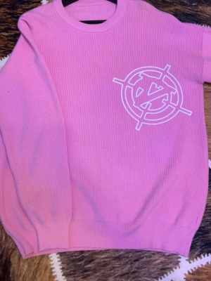 vicinity tröja - Säljer en rosa stickad tröja med ett vitt grafiskt tryck på bröstet. Tröjan har rund halsringning och ribbade muddar vid ärmslut och nederkant. Perfekt för dig som gillar färg och vill sticka ut lite extra.