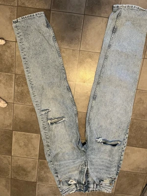 Ljusblå ripped mom jeans - Säljer ett par ljusblå mom jeans med slitningar på båda benen. Jeansen har hög midja, raka ben och klassisk femficksmodell. Tillverkade i denim med en cool vintagekänsla och trendiga hål vid knäna.