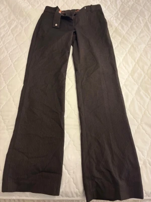 Bruna kostymbyxor med smala ränder - Snygga bruna kostymbyxor med diskreta smala kritstreck och raka, vida ben. Byxorna har bälteshällor och medföljande tygbälte. Perfekta för en stilren och trendig look. Materialet ser ut att vara en blandning med lite struktur.