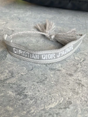 Grått Christian Dior tygarmband - Snyggt grått tygarmband från Christian Dior med broderad logga i vitt. Armbandet har justerbar knytning med tofsar i ändarna för en trendig look. Perfekt accessoar för att lyfta din outfit med en exklusiv touch.