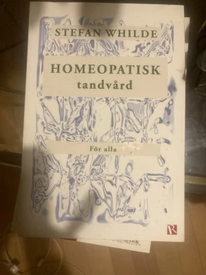 Homeopatisk tandvård av Stefan Whilde - Boken 'Homeopatisk tandvård' av Stefan Whilde utforskar alternativa metoder för tandvård med fokus på homeopati. Omslaget är vitt med abstrakta lila mönster och gröna textfält. Perfekt för dig som är nyfiken på holistisk hälsa och naturliga behandlingsmetoder.