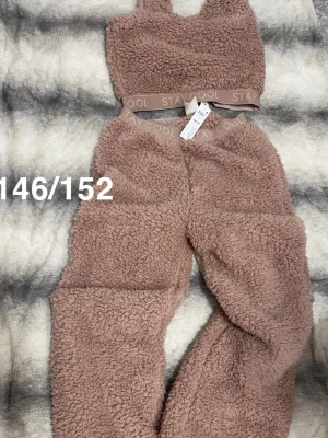 Rosa fluffigt loungewear-set - Mysigt loungewear-set i rosa teddyfleece med croppad topp och matchande byxor. Båda delarna har bred resår med texten 'STAY COOL'. Perfekt för chill dagar hemma. Storlek 146/152.🌸 Ny med lapp. 