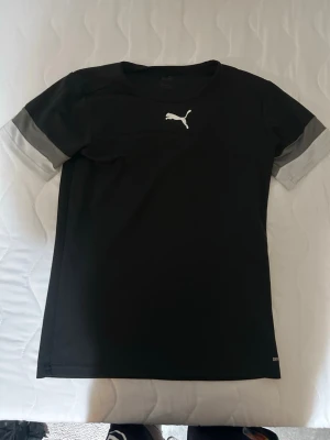 Svart tränings-tshirt från Puma - Snygg svart tränings-tshirt från Puma med grå detaljer på ärmarna och klassisk Puma-logga i vitt på bröstet. Tillverkad i ett lätt och snabbtorkande material som passar perfekt för sport och träning.