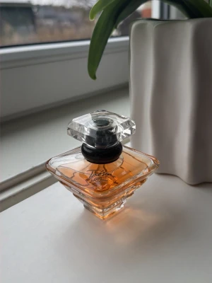 Lancôme Trésor Eau de Parfum - Säljer en elegant parfym från Lancôme, Trésor Eau de Parfum. Flaskan är fyrkantig med mjuka kanter och har en klar, kristalliknande kork. Vätskan är bärnstensfärgad och flaskan är tillverkad i glas. Perfekt för dig som gillar lyxiga dofter.