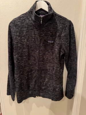 Patagonia full-zip fleece svart melange stl M herr - Patagonia full-zip i mjuk mikrofleece, svart/grå melange. Ståkrage, två handfickor. Gott skick. litet hål på ärmen (myrstort, se bild 6). Stl M herr.