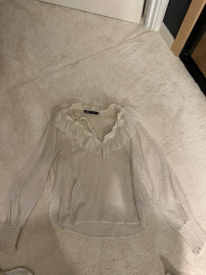 Vit blus från Zara - Söt vit blus från Zara med diskret prickigt mönster och volangdetaljer längs ringningen. Blusen har långa ärmar med breda smockade muddar och är gjord i ett tunt, transparent material som ger en romantisk känsla. Skriv för frågor osv🥰