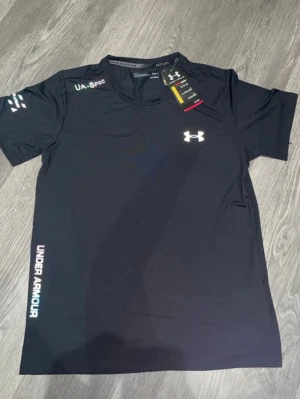 Svart tränings-tshirt Under Armour - Snygg svart tränings-tshirt från Under Armour med logga på bröstet och text längs sidan. Tillverkad i ett lätt och snabbtorkande funktionsmaterial, perfekt för gym eller löpning. Rund halsringning och korta ärmar för maximal rörelsefrihet.