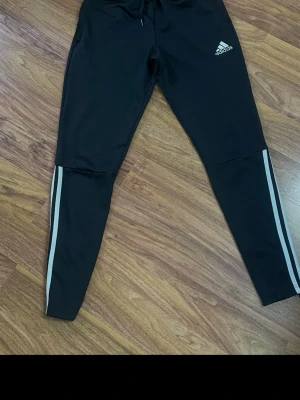 Svarta Adidas mjukisbyxor med ränder - Säljer ett par svarta mjukisbyxor från Adidas med vita ränder längs benen och dragsko i midjan. Byxorna har smal passform och Adidas-logga på låret. Perfekta för chill eller träning.