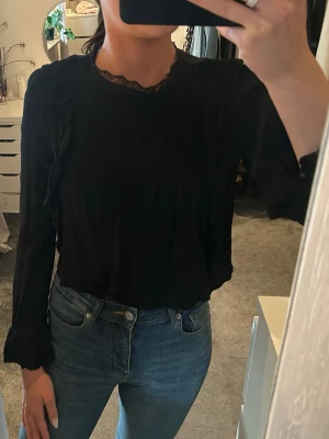 Svart blus med detaljer  - Snygg svart blus med lång ärm och fina volangdetaljer längs axlarna. Blusen har rund halsringning med spetskant och diskreta veck framtill. Perfekt att styla med jeans eller kjol 