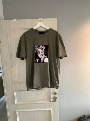 Olivgrön t-shirt Limitato x Ellen von Unwerth - Olivgrön t-shirt med rund hals och korta ärmar från Limitato x Ellen von Unwerth. Framsidan har ett svartvitt fotoprint med konstnärligt motiv. T-shirten är gjord i mjuk bomull. Dessvärre med liten missfärgning vid halsen, annars knappt använd. Storleken är L, men sitter som medium. Pris kan diskuteras, hör av dig vid minsta fråga!