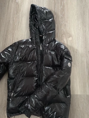 Svart glansig pufferjacka Moncler - Säljer en svart glansig pufferjacka från Moncler med huva och dragkedja framtill.  Perfekt för dig som vill ha både stil och värme under kalla dagar. Bra skick förutom att vänster framficka är lite trasig. Pris kan diskuteras vid snabb affär