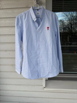 Blårandig skjorta från Ami Paris - Klassisk blårandig skjorta från Ami Paris med röd broderad logga på bröstet. Skjortan har lång ärm, knappar framtill och är tillverkad i mjuk bomull. Perfekt för en clean och stilren look. Säljer den då den inte passade i storlek, den är i XL men skulle säga att den passar L. Är 194 och den blev lite för kort.