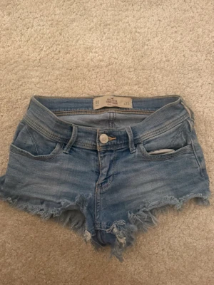 Blå jeansshorts från Hollister - Säljer ett par ljusblå jeansshorts från Hollister med fransiga kanter och slitningar. Klassisk femficksmodell med knapp och dragkedja framtill. Perfekta för sommaren och ger en avslappnad vibe.