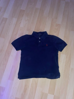 Marinblå polotopp från Polo Ralph Lauren - Säljer en klassisk marinblå polotopp från Polo Ralph Lauren med korta ärmar och krage. Toppens ikoniska röda logga är broderad på bröstet och den har knappar framtill. Perfekt för dig som gillar stilrena och tidlösa plagg.