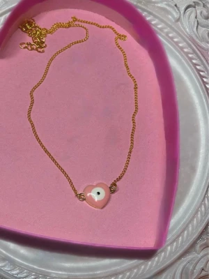 Guldhalsband med rosa hjärthänge/ondaögat - Sött halsband med guldfärgad kedja och ett rosa hjärta med ondaögat. Perfekt accessoar för att addera en gullig touch till din outfit. Hjärtat har en blank finish och kedjan är tunn och elegant.