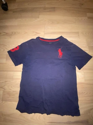 Blå Ralph Lauren t-shirt med röd logga - Mörkblå t-shirt från Ralph Lauren med röd broderad logga på bröstet och röd detalj på ärmen. Klassisk passform och rund halsringning med röd insida. Tillverkad i mjuk bomull, perfekt för en avslappnad stil. 