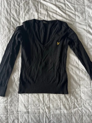 Svart stickad tröja från Lyle & Scott - Svart stickad tröja från Lyle & Scott med djup V-ringning och lång ärm. Tröjan har en gul broderad logga på bröstet och ribbade muddar vid ärmslut och nederkant. Perfekt för lager på lager och enkel att matcha.💗