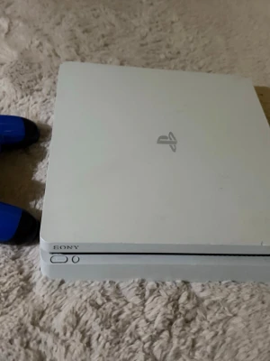 Vit Sony PlayStation 4 Slim konsol - Pris går att ändras. Säljer en vit PlayStation 4 Slim från Sony. Konsolen har en stilren, rektangulär design med rundade hörn och PlayStation-loggan på ovansidan. En blå handkontroll med svarta detaljer ingår. Perfekt för gaming och streaming. Sladdarna finns också såklart. Köpare står för frakt
