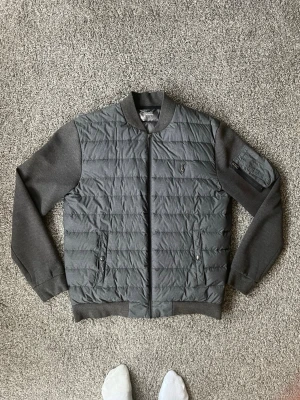 Grå cardigan från Polo Ralph Lauren - Snygg grå cardigan från Polo Ralph Lauren med quiltad front och släta ärmar. Jackan har ribbade muddar, dragkedja framtill, två fickor med tryckknapp och en ärmficka med dragkedja. Liten broderad logga på bröstet. Storleken är även M men sitter mer som S