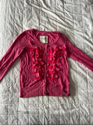 Rosa kofta med volang från Abercrombie - Söt rosa kofta från Abercrombie & Fitch med långa ärmar och knäppning framtill. Framsidan har flera volangdetaljer och en liten mörk logotyp nertill. Rund halsringning och ribbade muddar. Perfekt för dig som gillar färg och detaljer!💗