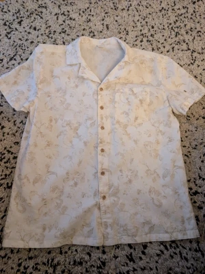 Vit blommig kortärmad linne skjorta stl 164 - Snygg vit beige skjorta med diskret blommigt mönster och korta ärmar. Skjortan har klassisk krage, bröstficka och knappar framtill. Perfekt för varma dagar och ger en avslappnad vibe.