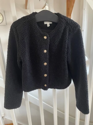 Svart jacka teddy med guldknappar - Mysig svart cardiganjacka i teddy-material med rund halsringning och långa ärmar. Jackan har sex dekorativa guldknappar framtill . Perfekt för lager på lager och ger en trendig look. Nyskick. 