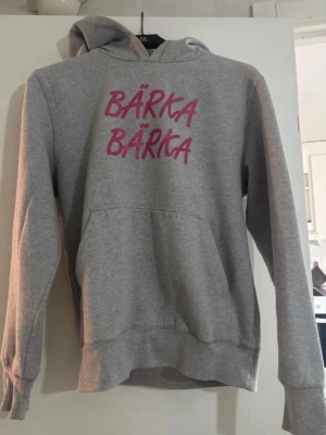 Grå hoodie med rosa text BÄRKA - Säljer en grå hoodie med huva och magficka. På bröstet står det 'BÄRKA BÄRKA' i rosa tryck. Hoodien har långa ärmar och ribbade muddar. Perfekt för dig som vill ha en chill och snygg stil.