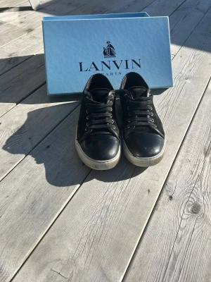 Svarta Lanvin sneakers med glans  - Snygga svarta sneakers från Lanvin i mocka med vita gummisulor och svarta skosnören. Klassisk låg modell med metallhål för snörning och diskret design. Perfekta för dig som gillar stilrena och exklusiva skor. Nypris runt 5000kr. Extrasnören följer med
