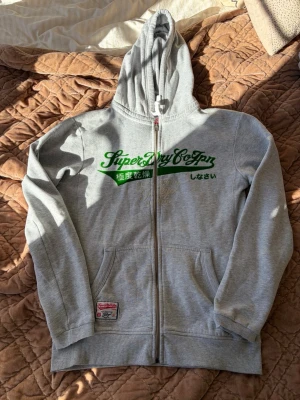 Superdry ziphoodie - Grå Superdry ziphoddie med grönt tryck på bröstet. 