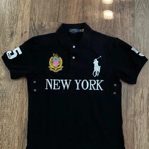 Chief Keef Polo Ralph Lauren t-shirt - Svart t-shirt från Polo Ralph Lauren med stor vit broderad logga, texten NEW YORK framtill och patchdetaljer på bröstet. Klassisk pikékrage och korta ärmar. Snygg kontrast mellan svart tyg och vita detaljer. Perfekt för dig som gillar streetstyle och sportig vibe.