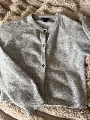 Ljusgrå kofta från Lindex - Mysig ljusgrå kofta från Lindex med rund halsringning och knäppning framtill. Koftan har långa ärmar, ribbade muddar och är tillverkad i ett mjukt, stickat material som känns skönt mot huden. Perfekt att slänga på sig när det är lite kyligt ute.