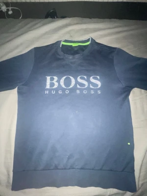 Hugo Boss Tröja  - Mörkblå sweatshirt från Hugo Boss med stort BOSS-tryck på bröstet och rund halsringning. Tröjan har ribbade muddar vid ärmslut och nederkant samt en diskret grön detalj vid kragen. Perfekt för en avslappnad och stilren look. Storleken på tröjan är M, men passar även till S. För mer info och bilder Dm😊