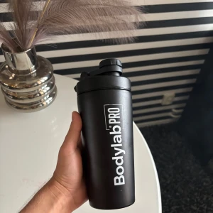 Svart BodylabPRO shaker - Säljer en svart shaker från BodylabPRO med vit logga och texten 'POWER YOUR PRO'. Tillverkad i plast och metall, med skruvlock och inbyggd mixer för smidiga proteinshakes. Perfekt för gymmet eller träningen.
