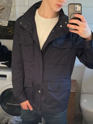 Field jacket - Snygg mörkblå field jacket med fyra stora fickor framtill och knappdetaljer. Jackan har hög krage, dragkedja och tryckknappar. Perfekt för dig som gillar en stilren och funktionell look. Passar till jeans och t-shirt för en avslappnad outfit.